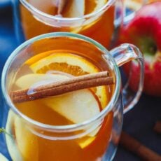 Two glass mugs of apple cider topped with apples, oranges, and cinnamon sticks