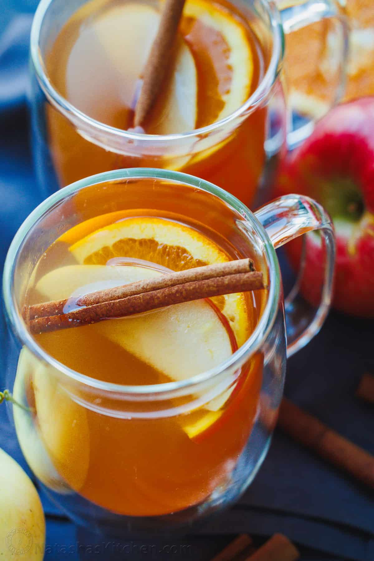 Two glass mugs of apple cider topped with apples, oranges, and cinnamon sticks