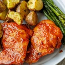 Two baked BBQ chicken thighs on a white plate next to roasted potatoes and asparagus.