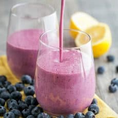 Two glass cups with a blueberry mango smoothie and blueberries and a halved lemon around the cups