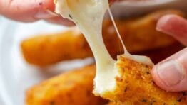 Two hands pulling apart a mozzarella stick with a plate of cheese sticks in the background.