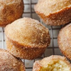Cinnamon muffins coated in cinnamon sugar on a wire rack, with one muffin on its side with a bite missing.