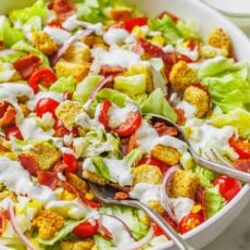 Close up of homemade garden salad in a large white bowl topped with croutons and drizzled with Ranch dressing, with two serving spoons.