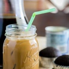 Iced coffee with condensed milk in a mason jar with a straw and cupcakes in the background
