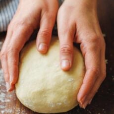 Two hands holding a ball of homemade pizza dough on a floured wooden countertop.