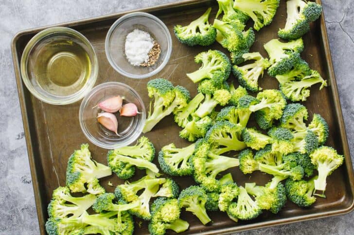 Raw broccoli florets with oil, garlic and seasonings in ramekins on a bakig sheet.