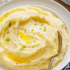 A bowl of sour cream mashed potatoes topped with melted butter and freshly chopped chives, with a spoon.